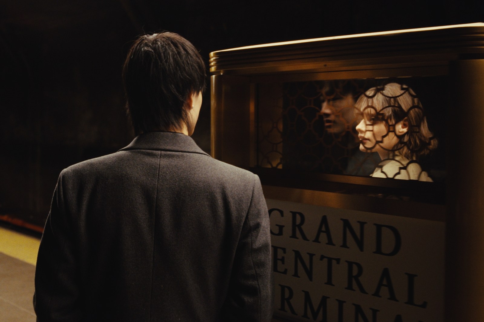 Film still: K stands before the Grand Central Terminal information booth as a woman's reflection layers over his in the brass grille